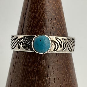 Può includere: Un anello d'argento con una pietra centrale turchese. La fascia presenta un motivo ripetuto a forma di onda. L'anello è esposto su un supporto di legno, evidenziando il suo design. Il turchese è di un colore blu vibrante.
