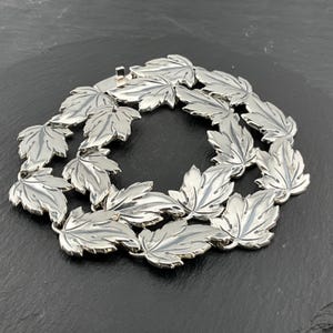 Puede incluir: Un collar plateado con un diseño de hojas repetidas. Las hojas están detalladas con vetas y conectadas para formar una forma circular. El collar tiene un cierre de broche y se muestra sobre una superficie oscura.