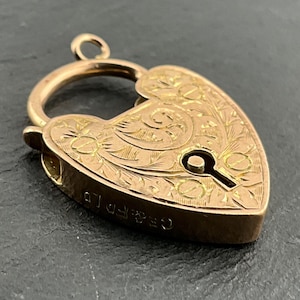 May include: A heart-shaped gold padlock charm featuring intricate floral engravings on the front surface. The metal piece includes a small circular bail at the top and the stamped text "G&F D" visible along the side edge of the gold casing.