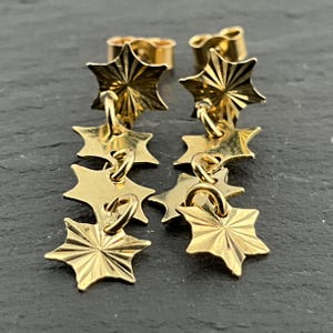 May include: A pair of gold-toned drop earrings featuring a vertical chain of four star-shaped charms. Each star has a polished finish with radiating etched lines emanating from the center. The earrings include standard post-style backings.