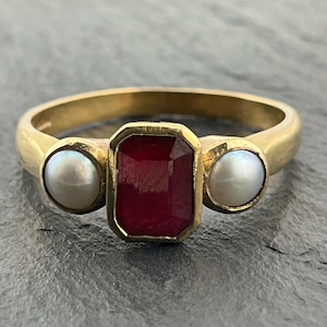 May include: A gold ring featuring a central rectangular, deep red gemstone set in a bezel, flanked by two round, white pearls on either side. The band is smooth and polished, showcasing a classic three-stone design with a warm yellow metal finish.