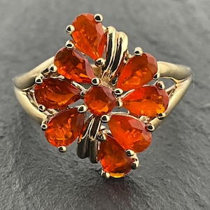 May include: A gold ring featuring a central circular orange gemstone surrounded by eight pear-shaped orange gemstones arranged in a floral pattern. The metal band is smooth and polished, providing a bright contrast to the vibrant orange stones.