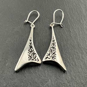 May include: A pair of silver dangle earrings with a triangular shape and a detailed, intricate design. The earrings have a delicate, openwork pattern that resembles branches or vines.