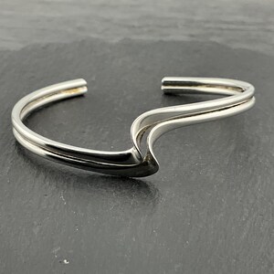 Könnte beinhalten: Ein silberfarbenes Armreif-Armband mit einem wellenförmigen Design. Das Armband hat eine glatte, polierte Oberfläche und eine Doppelbandstruktur. Das offene Design ermöglicht ein einfaches Tragen. Das Armband ist ein Schmuckstück.