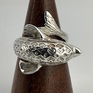 May include: A silver fish-shaped ring. The ring design features a fish with detailed scales and fins. The ring wraps around the finger, with the fish's body forming the main part of the ring.