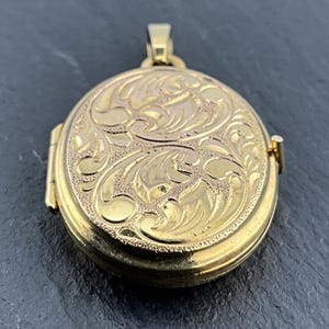 May include: An oval gold-coloured locket featuring an intricate engraved scrollwork pattern across the front surface. The locket includes a hinged side mechanism and a small top bail for attachment, showcasing a polished finish with textured details.
