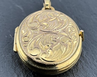 May include: An oval gold-toned locket featuring an intricate engraved scrollwork pattern across the front surface. The locket includes a hinged side mechanism and a small top bail for attachment, showcasing a polished finish with textured details.
