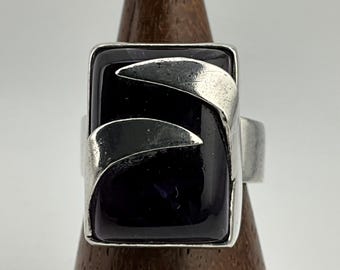 May include: A silver ring with a rectangular, dark purple stone. The ring features two curved silver accents on the face. The ring is displayed on a wooden stand against a white background.