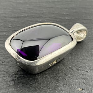 Puede incluir: Un colgante de plata con una piedra preciosa rectangular grande de color morado oscuro. El colgante tiene un diseño simple y elegante.