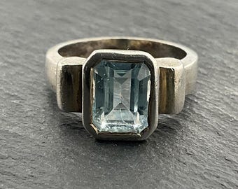 May include: A silver ring with a rectangular, light blue gemstone. The gemstone is set in a bezel setting, and the band has a simple, modern design. The ring is likely made of sterling silver.