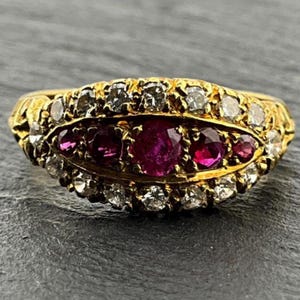 May include: A gold ring featuring a central row of five round-cut deep red rubies, surrounded by a halo of small, clear, round-cut diamonds. The band is crafted from yellow gold, with the stones set in a horizontal, elongated oval arrangement.