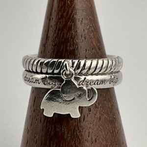 Puede incluir: Juego de anillos de plata con un anillo superior texturizado y un anillo grabado con "dream big". Un pequeño dije de elefante cuelga de los anillos. Los anillos se exhiben en un cono de madera.