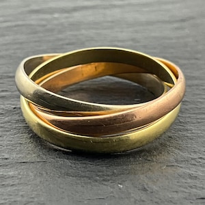 May include: A tri-colour rolling ring featuring three interlocking bands in yellow gold, rose gold, and white gold. The smooth, polished metal bands are intertwined to form a single circular piece of jewellery with a rounded profile and reflective finish.
