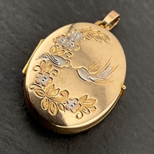 May include: Gold-tone oval locket with a floral and bird design. The locket features two birds in flight, with silver-tone accents, surrounded by flowers and leaves. The locket has a gold-tone bail for a chain.