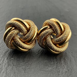 May include: A pair of gold-toned stud earrings featuring a knot design. Each earring is constructed from intertwined metal strands, alternating between smooth, polished surfaces and textured, cross-hatched sections that create a dimensional look.