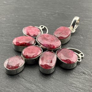 May include: A silver pendant with seven oval, deep red rubies. The rubies are set in individual bezels, arranged in a floral or starburst design. The pendant has a bail for a chain.