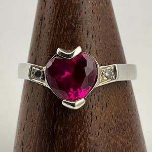 Op de afbeelding: Zilveren ring met een grote, gefacetteerde, hartvormige, dieprode edelsteen. De ring heeft kleine accentstenen aan weerszijden van het hart. Gepresenteerd op een houten standaard.