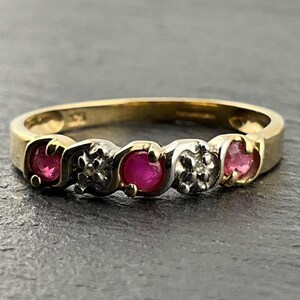 Könnte beinhalten: Ein Goldring mit drei runden roten Rubinen und zwei kleinen silberfarbenen floralen Fassungen mit eingelassenen klaren Steinen. Das Design wechselt zwischen den roten Rubinen und den floralen Akzenten und bildet ein durchgehendes Muster.