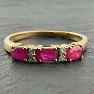 May include: A gold ring featuring three oval-cut red rubies alternating with two pairs of small round-cut clear diamonds. The stones are set in a horizontal row along the front of the polished gold band, creating a continuous, symmetrical design.