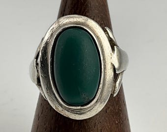 May include: A silver ring with an oval, dark green stone. The ring has a textured band and decorative elements on the sides. The stone is set in a bezel setting, and the ring is displayed on a wooden stand.