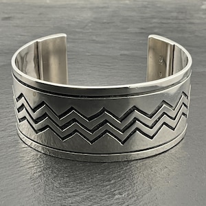 May include: A silver cuff bracelet with a geometric chevron pattern. The bracelet is made of polished metal and has a smooth, rounded surface.