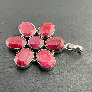 May include: A silver pendant with seven oval rubies, arranged in a floral design. The rubies are a rich red hue, set in individual silver bezels. The pendant includes a silver bail for attaching to a chain. The pendant is approximately 3 cm in diameter.