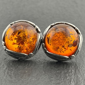 May include: A pair of round stud earrings featuring polished amber stones with inclusions. The amber is set in a silver-toned metal with a decorative leaf-like design around the amber stones. The earrings are displayed on a dark gray surface.