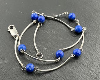 May include: A silver bracelet featuring vibrant blue gemstone beads. The bracelet has a delicate design with curved silver bars connecting the beads. The clasp is a simple, secure design. The bracelet is displayed on a dark, textured surface.