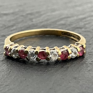 May include: A gold band ring featuring an alternating row of round-cut red and clear stones set in a prong setting. The metal band has a smooth finish and the stones are arranged in a repeating pattern across the front half of the jewelry piece.