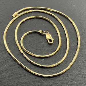 Op de afbeelding: Een goudkleurige slangenketting met een glad, flexibel schakelontwerp. De ketting is in een cirkelvormig patroon gewikkeld en bevat aan één uiteinde een veilige karabijnsluiting, wat zorgt voor een gepolijste en uniforme uitstraling.