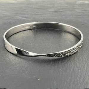 Könnte beinhalten: Ein silberfarbenes Armreif-Armband mit einem gedrehten Design. Eine Seite des Armbands hat ein strukturiertes, gepunktetes Muster. Das Armband ist kreisförmig und scheint aus Metall zu sein. Der Text "SODA 925N" und "85" ist auf dem Armband sichtbar.