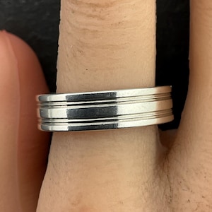 May include: A silver ring with three parallel, grooved bands. The ring is shown on a finger, with a close-up view highlighting the polished metal and the ring's design. The ring is a simple, yet elegant piece of jewelry.
