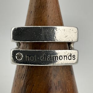 Peut inclure: Une bague en argent avec un design à double bande. La bande inférieure porte l'inscription gravée "hot diamonds". La bague est présentée sur un support en bois, mettant en valeur sa finition métallique polie.