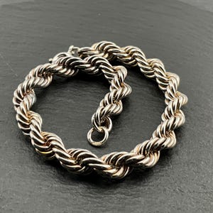 May include: A silver bracelet featuring a rope design. The bracelet is constructed from twisted metal strands, giving it a textured and detailed look. It has a simple hook and eye clasp. The bracelet is presented on a dark surface.