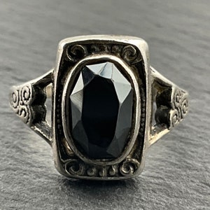 Puede incluir: Un anillo de plata con una piedra preciosa negra engastada en un bisel cuadrado. El anillo tiene un diseño decorativo en la banda.