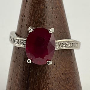 May include: A silver ring with a large, oval-cut, deep red gemstone. The band is embellished with small, clear stones on either side of the central stone. The ring is displayed on a wooden stand.