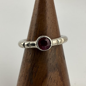 May include: A silver ring with a round, deep red gemstone at the center. The band has a series of small, rectangular cutouts. The ring is displayed on a wooden stand against a white background.