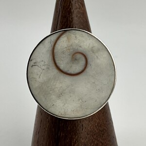 May include: A large, round ring with a silver band. The ring's face features a light grey, circular stone with a brown spiral design. The ring is displayed on a wooden stand.