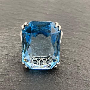 Può includere: Un anello con una grande pietra preziosa blu rettangolare, con montatura in argento. La pietra è di un blu vibrante e trasparente, con molteplici sfaccettature. La montatura dell'anello presenta dettagli decorativi argentati. L'anello è esposto su una superficie grigio scuro.