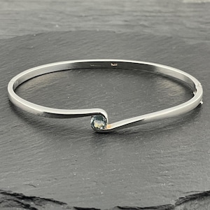 May include: A silver-toned bangle bracelet with a single, round, light blue gemstone. The bracelet has a simple, elegant design with a curved section holding the gemstone. The clasp is visible on the right side.