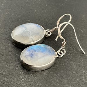 May include: A pair of silver-toned drop earrings. Each earring features an oval, iridescent moonstone set in a silver bezel. The moonstones display a mix of white, blue, and gray hues. The earrings have a hook closure.