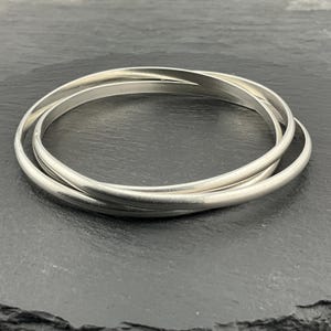 May include: Three interlocking silver bangles. The bangles have a smooth, rounded design and a subtle matte finish. They are arranged in a circular pattern, creating a layered effect. The jewellery is displayed on a dark, textured surface.
