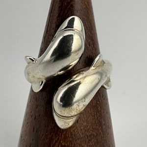 May include: A silver ring featuring two intertwined fish, each with a detailed, realistic design. The ring is displayed on a wooden ring holder.
