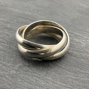 Pode incluir: Um anel prateado interligado com três faixas. As faixas são lisas e arredondadas, criando um design contínuo. O anel é exibido em uma superfície cinza escuro, destacando seu brilho metálico. O anel parece ser feito de prata esterlina.