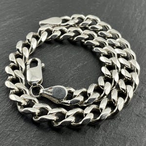 May include: A silver curb chain bracelet with a lobster clasp. The bracelet is made of thick, interlocking links. The metal has a bright, polished finish. The bracelet is shown against a dark, textured background.