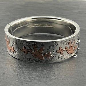 May include: Silver hinged bangle bracelet with etched floral and bird design. The birds are a copper colour. The bracelet is on a dark grey surface.