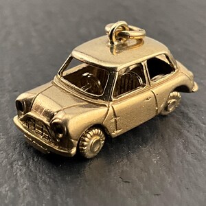 Może przedstawiać: Złoty, miniaturowy charms w kształcie samochodu z detalami, w tym kołami, oknami i dachem. Charms ma pętlę na górze do mocowania. Samochód ma klasyczny design, prawdopodobnie w stylu vintage lub retro.