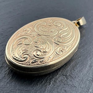 May include: An oval gold locket pendant featuring an intricate embossed scrollwork pattern across the front surface. The metal piece has a polished finish and includes a small, plain gold bail at the top for attachment to a chain or necklace.