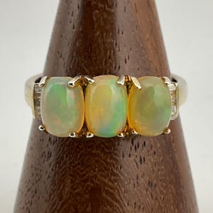 Puede incluir: Un anillo dorado con tres ópalos ovalados, cada uno con un juego de colores, engastados en una montura de garras. El anillo tiene pequeñas piedras de acento a cada lado de los ópalos. Los ópalos son de color amarillo y verde.