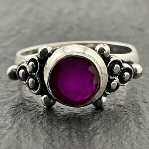 May include: A silver ring featuring a round, faceted, deep magenta gemstone set in the centre. The metal band displays an oxidised finish with decorative, raised spherical and scroll-like details flanking the central stone setting.